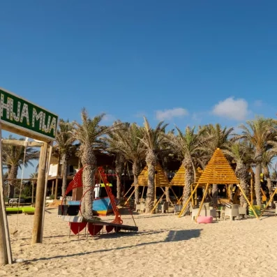 Plage sableuse avec cabanes en bois, palmiers et installation colorée sous un ciel bleu.