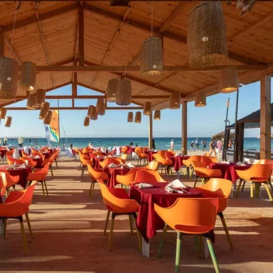 Restaurant en bord de mer avec chaises orange, tables dressées et vue sur la plage.