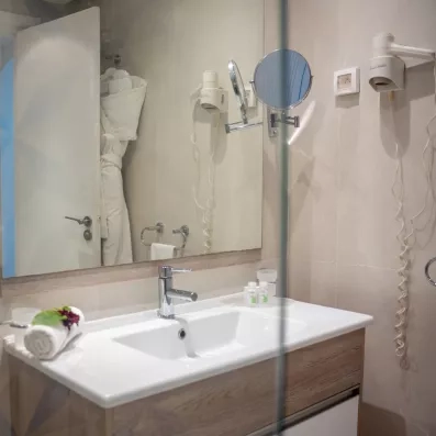 Salle de bain moderne de l'hôtel Ulysse à Djerba avec grand miroir, vasque blanche et produits d'accueil.