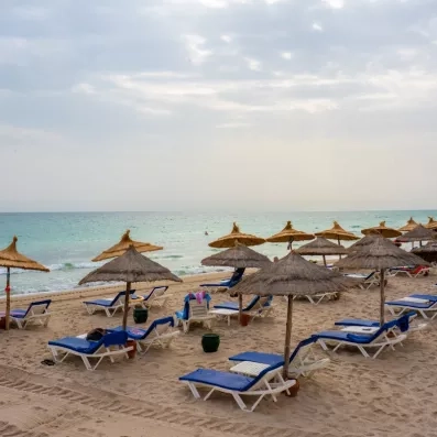 Plage de sable fin et parasols en paille à l'hôtel Ulysse Djerba au bord de l'eau turquoise.