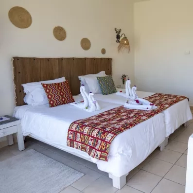 Chambre avec deux lits jumeaux décorés de serviettes en cygnes, coussins colorés et décorations murales en fibres naturelles.