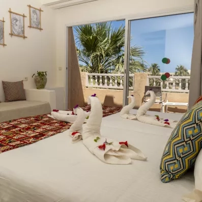 Chambre d’hôtel lumineuse avec lit décoré de serviettes en forme de cygnes, pétales de fleurs et vue sur palmiers depuis le balcon.