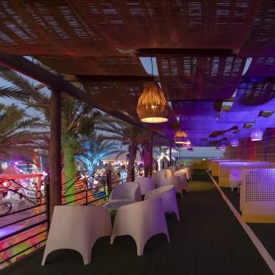 Lounge extérieur nocturne avec chaises blanches, éclairage coloré et ambiance tropicale chaleureuse.