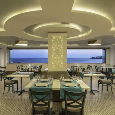 Une salle de restaurant intérieure élégante face à une large baie vitrée ouvrant sur la mer.