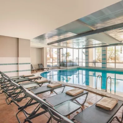 Une rangée de transats installés le long d'une piscine intérieure aux larges baies vitrées.