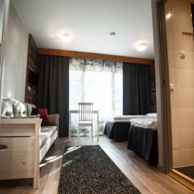 chambre lumineuse et moderne, aménagée avec deux lits simples, un petit coin table, un canapé clair et une salle de bain attenante, dans une ambiance chaleureuse mêlant bois et tons neutres.