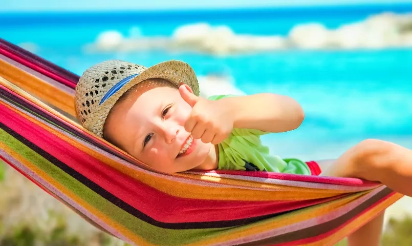 Un petit garçon avec un chapeau allongé dans un hamac coloré et montrant son pouce, en fond la mer turquoise